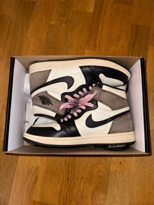 Air Jordan 1 High Mocha med rosa snören - Säljer ett par Air Jordan 1 High Mocha sneakers med svart, vit och brun mocka samt rosa skosnören i storlek 41. Klassisk Nike Swoosh på sidan och Air Jordan-logga vid ankeln. Skorna har en hög siluett och är riktigt snygga till streetwear-stilen.