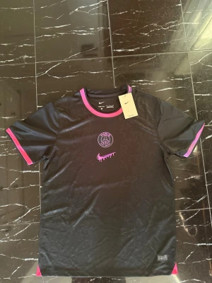 Svart PSG x Nike Dri-FIT t-shirt - Snygg svart PSG t-shirt från Nike med Dri-FIT-material. Tröjan har rosa och lila detaljer på krage och ärmslut, samt PSG-logga och en unik rosa Nike swoosh på bröstet. Perfekt för träning eller streetwear.