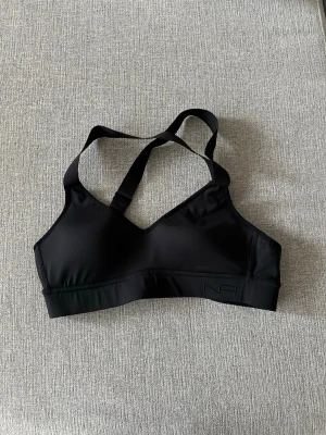 Svart sport-bh med korsade band - Snygg svart sport-bh med korsade axelband och justerbar knäppning bak. Bh:n har en enkel och stilren design utan vaddering och är tillverkad i ett stretchigt material som ger bra stöd. Perfekt för träning eller en aktiv vardag. Endast använd 2ggr!