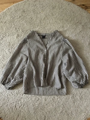 Randig blus med puffärm - Säljer en svartvit randig blus från Lindex med v-ringning och knäppning framtill. Blusen har långa puffärmar och är gjord i ett lätt, vävt material som känns luftigt. Perfekt till jeans eller kjol.