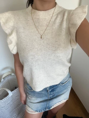 Vit stickad topp med volangärmar - Supersöt vit stickad topp med volangdetaljer på ärmarna och ribbad halsringning. Perfekt att matcha med jeans eller kjol för en trendig look. Toppens material är mjukt och känns lyxigt mot huden.