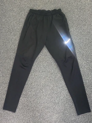 Nike Miller dri fit byxor  - Säljer ett par svarta Nike byxor med klassisk vit logga på låret. De har elastisk midja och är tillverkade i ett stretchigt syntetmaterial som passar perfekt för träning. Tight passform och stilren design.
