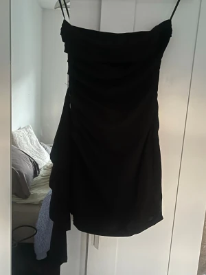 Svart strappless klänning med drapering - Säljer en svart, kort klänning med snygg drapering på sidan. Klänningen har en asymmetrisk detalj och är figurnära i modellen. Perfekt för dig som gillar en stilren och trendig look.