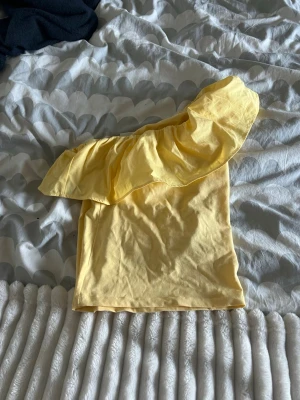 Gul oneshoulder topp med volang - Snygg gul topp i oneshoulder-modell med stor volang längs axeln. Perfekt för dig som vill sticka ut med en färgglad och trendig look. Tillverkad i mjukt bomullstyg som känns skönt mot huden.