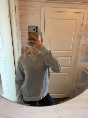 Grå sweatshirt från Ralph Lauren - Säljer en klassisk grå sweatshirt från Ralph Lauren med broderad logga på bröstet. Tröjan har rund halsringning, ribbade muddar vid ärmslut och nederkant samt en relaxed fit. Perfekt för en chill och stilren look.