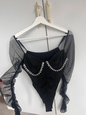 Svart meshbody med pärldetaljer - Snygg svart body med transparenta meshärmar och dekorativa pärlor längs bysten. Kroppsnära passform och off shoulder-design ger en trendig look. Materialet är en mix av stretchigt tyg och mesh. Köpt från shein, hittar dock inte nypris på den. Storlek S. Ena pärlbandet har släppt men jag har sen tidigare fixat det med en säkerhetsnål.
