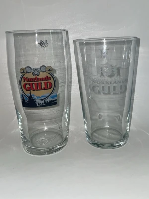 Norrlands Guld ölglas - 80kr glas  ölglas från Norrlands Guld, ett med färgat tryck och ett med frostad logga. Båda glasen är genomskinliga och har klassisk pintform. Perfekta för dig som gillar att samla på ölglas eller vill ha snygga glas till festen.