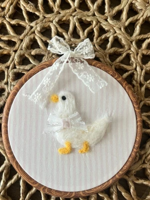 Broderad tavla med anka och spetsrosett - Söt handgjord broderad tavla med en vit anka i ull, dekorerad med gul näbb och ben samt en spetsrosett runt halsen. Tavlan har en rund träram och en extra spetsrosett upptill. Perfekt som gullig inredningsdetalj.