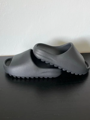 Yeezy Slides - Svart - Svarta Yeezy Slides med mjuk och bekväm känsla. Den breda remmen sitter stabilt på foten och sulan ger bra grepp för både inne och ute.