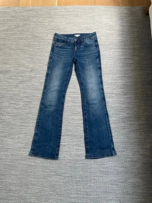 Blå bootcut jeans med broderade fickor - Säljer ett par blå bootcut jeans med broderade detaljer på bakfickorna. Jeansen har klassisk femficksmodell och stängs med knapp och dragkedja.