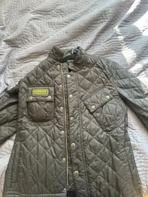 Svart quiltad jacka från Barbour - Snygg svart quiltad jacka från Barbour International med tryckknappar, dragkedja och flera fickor. Klassisk design med Barbour-logga på bröstet och detaljer i silver. Perfekt för dig som vill ha en fin och tidlös jacka. Pris kan diskuteras.