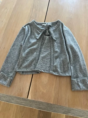 Grå cardigan med knyt från lager 157 - Grå mjuk cardigan från lager  157 med knytband i halsen och långa ärmar. Kortare modell med breda muddar vid ärmsluten. Perfekt att slänga över en topp för en chill och stilren look.