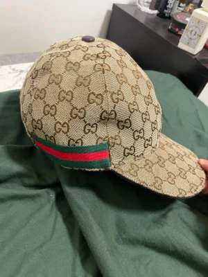 Beige keps från med GG-mönster - Säljer en beige keps med det klassiska GG-monogrammet över hela kepsen. På sidan finns en grön och röd rand som ger en ikonisk touch. Kepsen har böjd skärm och är tillverkad i canvasmaterial.