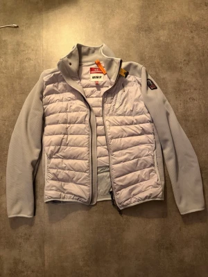 Ljusgrå dunjacka från Parajumpers - Säljer en ljusgrå dunjacka från Parajumpers med quiltad front och släta ärmar. Jackan har hög krage, dragkedja framtill och en ikonisk patch på ärmen. Materialet är en mix av polyester och syntet, perfekt för kalla dagar.om du brukar bära L så passar den dig perfekt