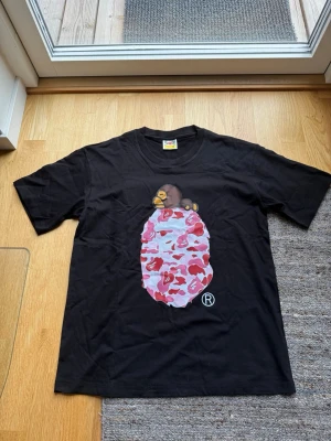 Svart BAPE t-shirt med rosa camo - Säljer en svart t-shirt från BAPE med ikoniskt Baby Milo-tryck på bröstet. Motivet har rosa, röd och vit camouflage och en apa ovanpå. Klassisk passform och rund hals. Perfekt för dig som gillar streetwear och vill sticka ut.