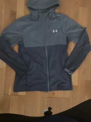 Gråblå vindjacka från Under Armour - Snygg tvåfärgad vindjacka från Under Armour i grått och mörkblått. Jackan har huva, hel dragkedja framtill och logga på bröstet. Tillverkad i lätt och tåligt material, perfekt för blåsiga dagar. Diskret tryck på ärmen och praktiska fickor med dragkedja.