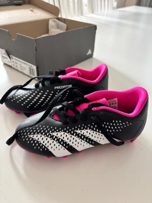  Predator svart/vit/rosa fotbollsskor - Snygga addida Predator fotbollsskor i svart med vita detaljer och rosa insida samt dobbar. Skorna har snörning och är tillverkade i syntetmaterial för bra grepp och kontroll på planen. Perfekta för dig som vill sticka ut med stil på matchen.