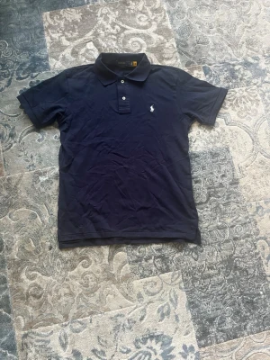 Marinblå pikétröja från Ralph Lauren storlek M - Klassisk marinblå pikétröja från Ralph Lauren med vit broderad logga på bröstet. Tröjan har krage, två knappar framtill och korta ärmar. Tillverkad i mjuk bomull som känns skön mot huden. Perfekt till jeans eller shorts för en clean look. Tröjan är helt ny och aldrig buren och har storlek M