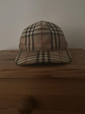 Beige Burberry keps med rutigt mönster - Säljer en klassisk beige keps från Burberry med det ikoniska rutiga mönstret i svart, vitt och rött. Kepsen har böjd skärm och justerbar rem bak. Perfekt accessoar för dig som vill ha en tidlös och trendig look och ännu perfekt inför sommaren 