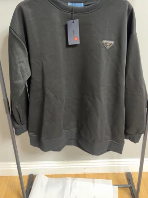 Svart sweatshirt från Prada - Snygg svart sweatshirt från Prada med det ikoniska trekantiga metall-logotypen på bröstet. Tröjan har rund halsringning, ribbade muddar vid ärmslut och nederkant samt en clean och stilren look. Tillverkad i mjukt bomullsmaterial som känns skönt mot huden.