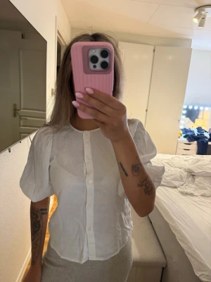 Vit puffärmad blus med knappar - Säljer en vit blus med puffiga korta ärmar och knäppning framtill. Blusen har en rak passform och är tillverkad i ett tunt, lätt material som ger en luftig känsla. Perfekt för en stilren och trendig look. Storlek xs men skulle säga passa xs-s. Aldrig använd så tröjan är i fint skick. 