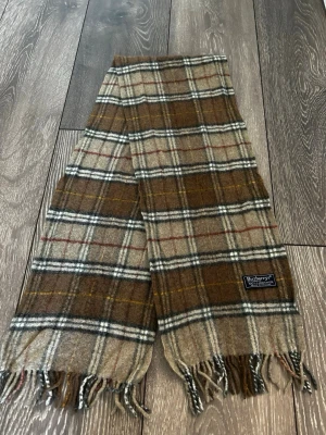 Rutig halsduk från Burberrys i ull - Klassisk rutig halsduk från Burberrys i 100% lammull. Färgerna går i brunt, beige, vitt och inslag av rött och gult. Halsduken har fransar i ändarna och den ikoniska Burberrys-loggan påsydd. Perfekt accessoar för att lyfta din outfit.