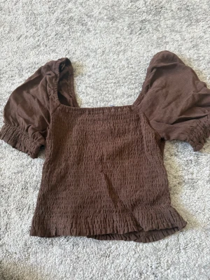 Brun puffärm topp från KIABI - Säljer en brun topp från KIABI med puffärmar och smockad struktur. Toppen har fyrkantig ringning och korta ärmar med resår vid ärmslut. Perfekt för dig som gillar trendiga detaljer och en bekväm passform.