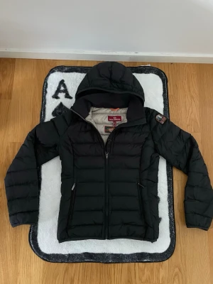 Svart dunjacka från Parajumpers - Snygg svart dunjacka från Parajumpers med huva och dragkedja framtill. Jackan har två fickor med dragkedja och en patch med logga på ärmen. Perfekt för kalla vinterdagar och riktigt stilren design.