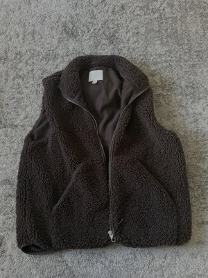 Brun teddyväst från H&M - Mysig brun teddyväst från H&M med dragkedja framtill och två stora fickor. Västen har en hög krage och är gjord i ett fluffigt, mjukt material som ger en avslappnad och trendig look. Perfekt att slänga över en hoodie eller tröja för extra värme.