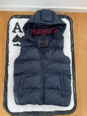 Marinblå dunväst från Tommy Hilfiger - Snygg marinblå dunväst från Tommy Hilfiger med huva och dragkedja. 