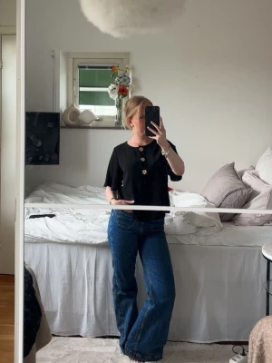 Svart topp med stora knappar - Säljer en svart topp med korta ärmar och tre stora dekorativa knappar framtill. Toppen har en rak, lite loose passform och är tillverkad i ett mjukt bomullstyg. Perfekt att styla med jeans eller kjol för en enkel men snygg look.