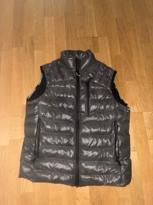 Grå dunväst  - Snygg grå dunväst med hög krage och svarta detaljer. Västen har tre dragkedjeförsedda fickor framtill och quiltad design. Materialet är glansigt och lätt, perfekt för lager-på-lager. Diskret logga på insidan och dragkedjor i svart.