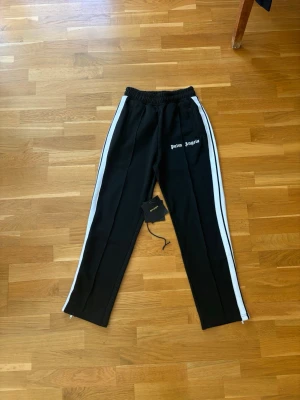 Palm Angels svarta mjukisbyxor - Palm Angels svarta mjukisbyxor med vita sidoränder och logga broderad på vänster sida. Byxorna har elastisk midja, raka ben och dragkedjor vid benslut. Tillverkade i ett mjukt och bekvämt material, perfekta för en avslappnad och trendig look.