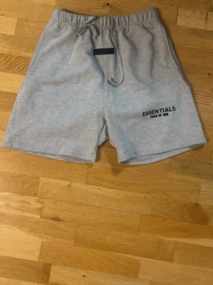 Grå shorts från Essentials Fear of God - Snygga grå shorts från Essentials Fear of God med elastisk midja och snörning. Mjukt bomullsmaterial och logga tryckt på ena benet. Perfekta för en avslappnad och trendig stil.