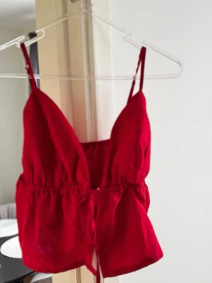 Röd linnetopp med smala axelband - Snygg röd linnetopp med smala justerbara axelband och v-ringad. Toppen har en croppad passform och knytning i midjan för extra detalj. Perfekt för varma dagar och somriga outfits. XS men passar även S