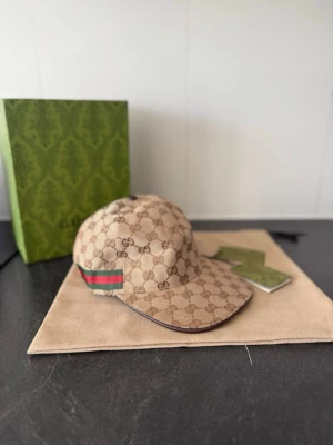 Beige keps från Gucci med GG-mönster - Snygg beige keps från Gucci med det klassiska GG-monogrammönstret över hela kepsen. På sidan finns den ikoniska gröna och röda randen. Kepsen har böjd skärm och justerbar rem bak. Materialet är canvas med läderdetaljer.