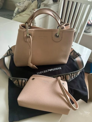 Ljusrosa handväska Emporio Armani - Snygg ljusrosa handväska från Emporio Armani i strukturerat läder. Väskan har silverfärgade metalldetaljer, dubbla handtag och avtagbar axelrem med logga. Tillhörande mindre clutch i samma färg och material ingår. Slitage : alla 4 hörn har spår av användning (se sista bild )