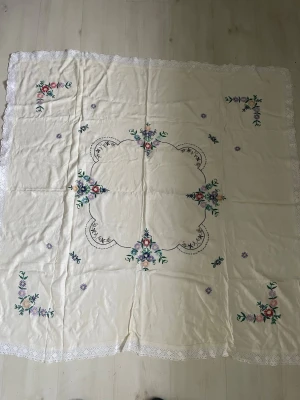 Gammal Bordduk med broderade blommor - Stor, ljus bordduk med broderade blommor i rosa, lila och grönt. Duken har spetskant runt om och ett klassiskt, romantiskt mönster med blommor i hörnen och i mitten. Perfekt för att ge ett vintage-inspirerat intryck till matbordet med spets runt om 