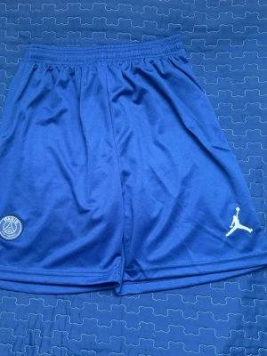 Blå PSG x Jordan shorts - Säljer ett par blå PSG x Jordan shorts med elastisk midja. Shortsen har PSG-logga på ena benet och Jumpman-logga på det andra. Tillverkade i ett lätt och ventilerande syntetmaterial, perfekta för fotboll eller träning.