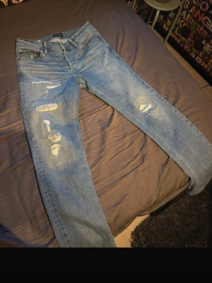 Ljusblå slitna jeans med raka ben - Säljer ett par ljusblå jeans med raka ben och slitna detaljer framtill. Jeansen har klassisk femficksmodell och är tillverkade i denim. Perfekta för en avslappnad och trendig look.