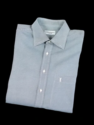 Randig skjorta från Yves Saint Laurent - Klassisk skjorta från Yves Saint Laurent i tunn bomull med smala blå och vita ränder. Skjortan har lång ärm, bröstficka med broderad logga och vita knappar framtill. Perfekt för en clean och stilren look.
