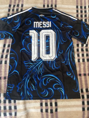 Argentina Messi tröja svart/blå Adidas - Snygg svart fotbollströja från Adidas med blått mönster, nummer 10 och Messi på ryggen. Framsidan har FIFA-märke och AFA-logga. Korta ärmar och klassiska vita ränder på axlarna. Perfekt för dig som älskar fotboll och vill sticka ut på planen. Helt ny med tagg. Priset är ej hugget i sten!