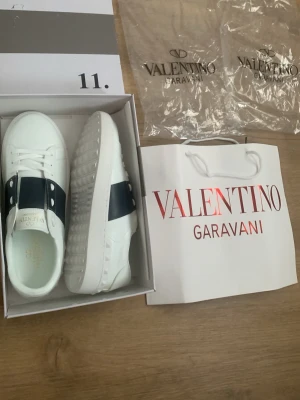 Valentino Garavani vita sneakers - Säljer ett par snygga vita sneakers från Valentino Garavani med bred svart rem över mitten och vita snören. Skorna har en chunky sula med nitar längs kanten och är tillverkade i slätt skinn. Perfekta för dig som gillar exklusiv och stilren design.