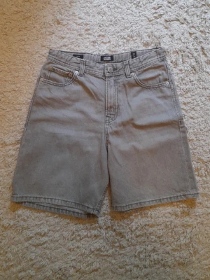 Grå denimshorts från Jack & Jones - Säljer ett par grå denimshorts från Jack & Jones med klassisk femficksdesign och knappstängning. Modellen är loose fit och har en enkel, stilren look som passar perfekt till sommaren. Materialet är bomull och shortsen har normal passning.