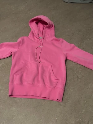 Rosa hoodie från Nike med huva - Säljer en snygg rosa hoodie från Nike med klassisk vit logga broderad på bröstet. Hoodien har huva med dragsko och en stor magficka framtill. Tillverkad i mjukt bomullsmaterial som är skönt att ha på sig. Perfekt för dig som gillar streetwear och sportig stil.
