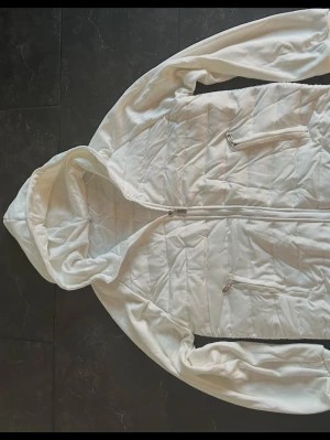 Vit quiltad hoodie med dragkedja - Säljer en clean vit hoodie med quiltat mönster och dragkedja framtill. Hoodien har två fickor med dragkedja och en stor huva. Perfekt för dig som gillar en stilren look och vill ha något bekvämt och snyggt. Materialet känns mjukt och skönt.