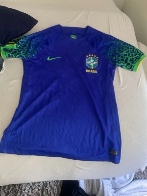 Blå Brasilien Nike fotbollströja - Snygg blå fotbollströja från Nike med Brasilien-emblem på bröstet och grönt Nike-märke. Ärmarna har ett unikt mönster i grönt och blått med leopardliknande detaljer. Perfekt för dig som älskar fotboll och vill sticka ut på planen.