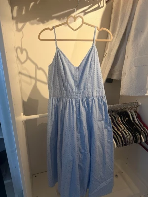 Ljusblå prickig klänning H&M - Söt ljusblå klänning från H&M med tunna axelband och v-ringning. Klänningen har ett prickigt mönster i mörkblått och en luftig, något utställd passform. Perfekt för varma dagar och enkel att styla med sneakers eller sandaler.