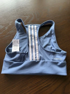 Blå sport-bh från Adidas - Snygg blå sport-bh från Adidas med vita detaljer och klassiska tre ränder på ryggen. Tillverkad i funktionsmaterialet Aeroready som andas och har meshpanel bak för extra ventilation. Perfekt för träning och sportiga aktiviteter.