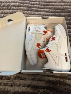 Air Jordan 4 Retro White/Orange - Säljer ett par Air Jordan 4 Retro sneakers i vitt läder med meshdetaljer och orange metallic-accenter. Klassisk siluett med synlig Air-enhet i sulan och ikonisk Jumpman-logga på plösen. Skorna har vita skosnören och en chunky sula för extra streetkänsla.